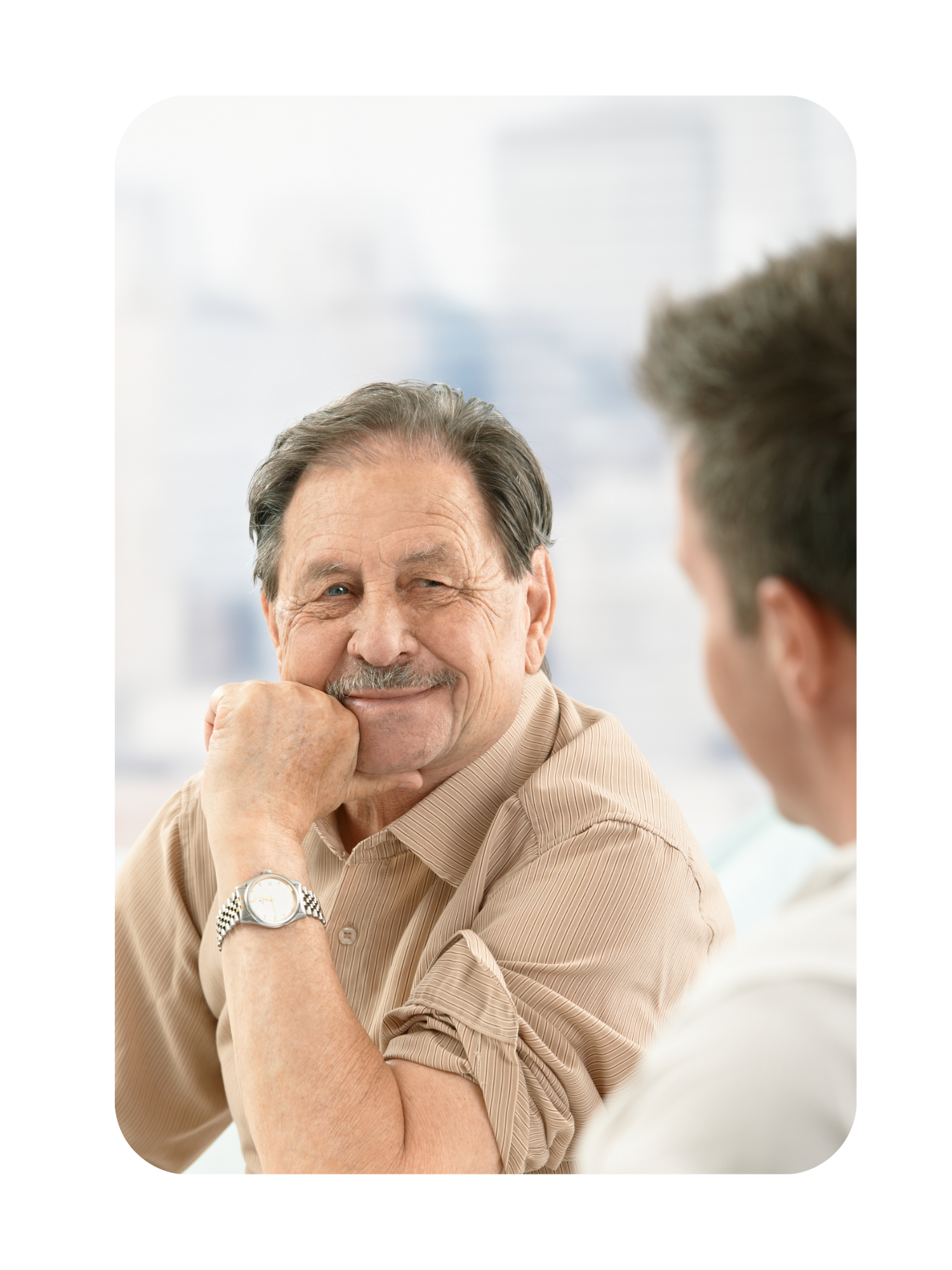 Male hipe replacement patient in a beige shirt sitting and smiling, with an orthopedic surgeon partially visible talking to him.