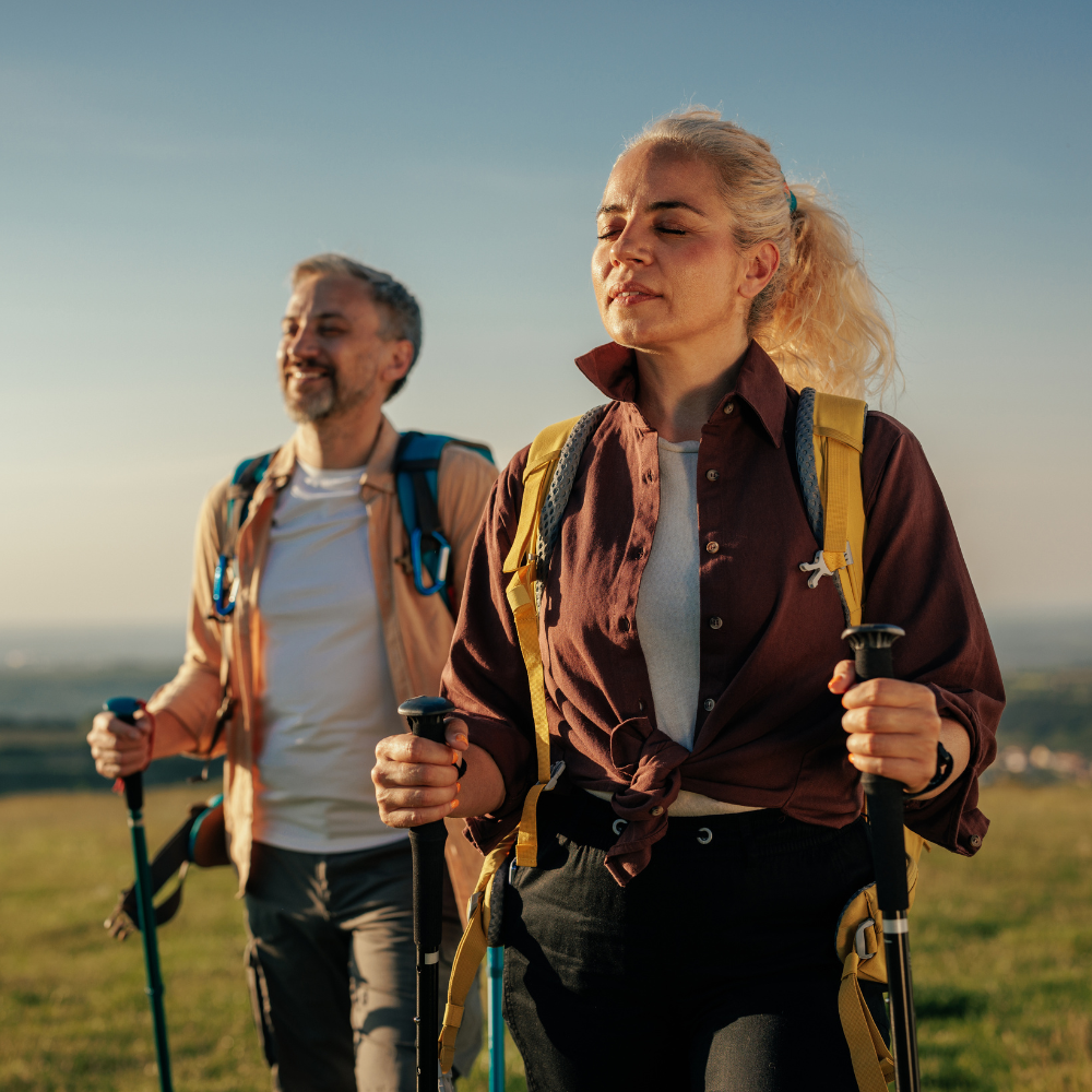 Two joint replacement patients with backpacks and walking sticks on a grassy field.