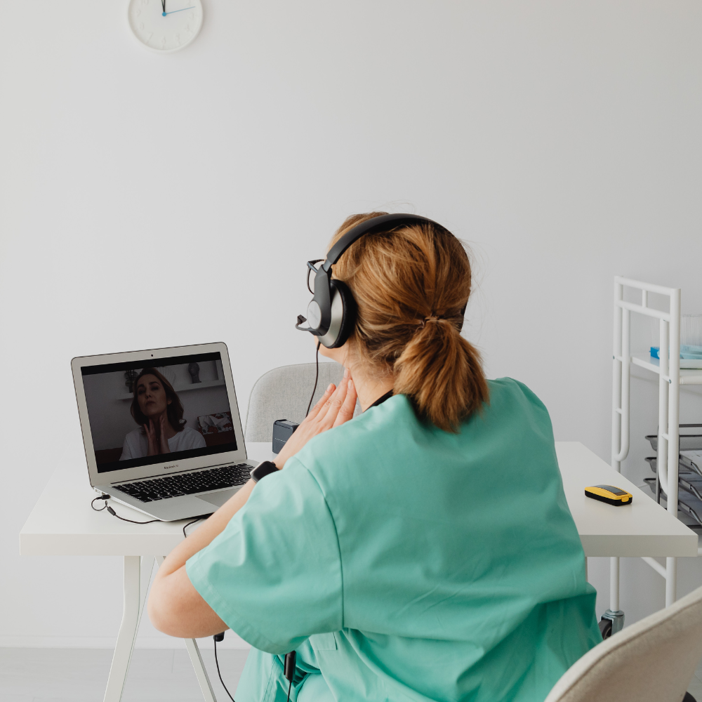 SurgicalRx health coach in teal shirt with headphones sitting at a laptop in a room with a white wall and clock.